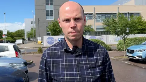 Dan Freeman is bald and looking straight down the camera. He is standing in a carpark at the Churchill Hospital in Oxford.