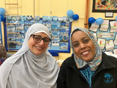 Two women wearing light coloured hijabs are smiling at the camera in front of an information board