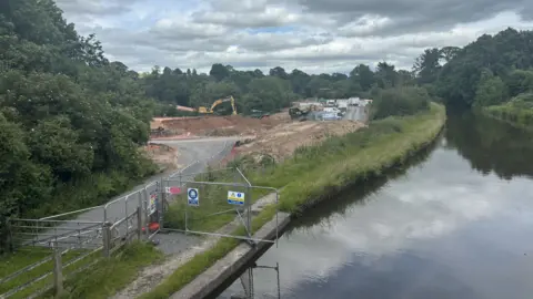 BBC/Chloe Hughes A canal is no the right hand side. On the left there is a gravel road leading up to a huge mound of earth that slops down in a drop. There is a yellow digger in the background and various other construction vehicles in the background