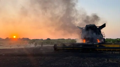 Cambridgeshire Fire and Rescue Service A large field that has been burnt with a combine harvester on fire, with two firefighters to the left. Trees are in the distance, and a setting sun. Smoke is billowing up from the combine harvester.