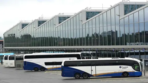 A number of blue, white and gold Translink Goldliner busses are lined up outside Belfast Grand Central Station. 
The large station can be seen in the background with a number of windows visible. 