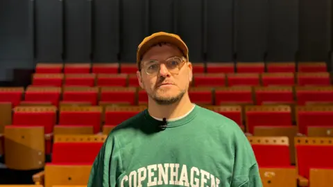 A head and shoulders shot of Paul Smith looking at the camera. He is wearing glasses, an orange cap and a green sweater with "Copenhagen" in white bold capital letters printed on the front. He is stood in front of red theatre chairs. The walls are black.