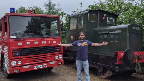 Lawrie Rose Lawrie Rose is pictured standing inbetween a green locomotive and red fire engine parked next to each other. He has long dark hair that is tied up and a dark beard. He is wearing a blue t-shirt with blue jeans. He has spread his arms out wide either side of him toward the vehicles. 