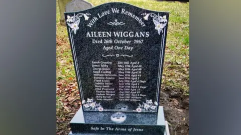 LDRS The gravestone inn the cemetery showing all the names of the babies, children and adults that are buried there. It is black marble with flowers in each corner. Aileen Wiggans name is at the top and the base says 'safe in the arms of Jesus'. 