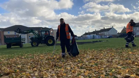Two workers in orange hi-vis jackets are litter picking among leaves on a Hartlepool field. A green tractor is in the background putting rubbish in a large trailer.