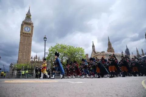 PA Media A low-down camera angle shows the military procession marching in Parliament Square outside Big Ben and the Houses of Parliament against a cloudy sky.