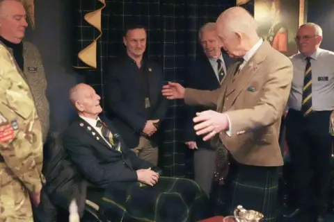 PA Media Jim Glennie, 99-year-old man wearing a striped tie and a jacket, speaking to King Charles in a light brown jacket and kilt, with his arms outstretched