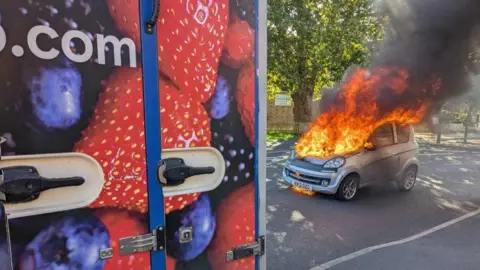 Ben Rowe The back of a delivery lorry with pictures of fruit on the side and a small silver car with flames coming from it with a tree in the background