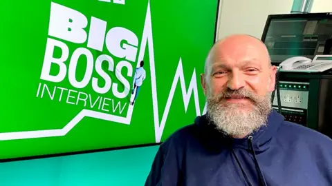 BBC Chris O'Shea, boss of Centrica, in the BBC's Big Boss studio. He has a big bushy beard and is wearing a blue hoody.