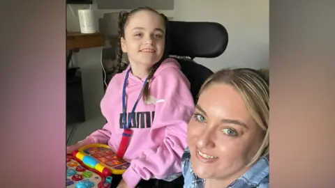 Family A girl in a pink Puma sweatshirt with plaited brown hair sits in a supportive therapy chair with an activity toy on her lap. A woman with blonde hair and a blue shirt is crouching or sitting next to her and looking up at the camera.