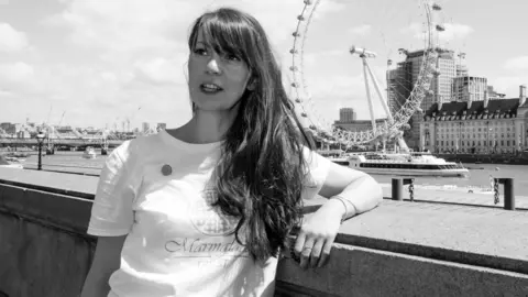 The Marmalade Trust Black and white image of Amy Perrin OBE in a white t-shirt with the words "Marmalade Trust" on the front. The Thames river and London Eye are in the background.