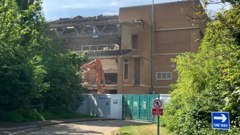 Shariqua Ahmed/ BBC Regional Pool building with a collapsed roof and frontage and an orange digger in front of it.