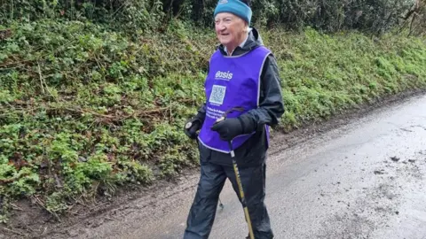 Derrick Downs aged 90 walking along a muddy path holding walking poles and wearing a mauve bib