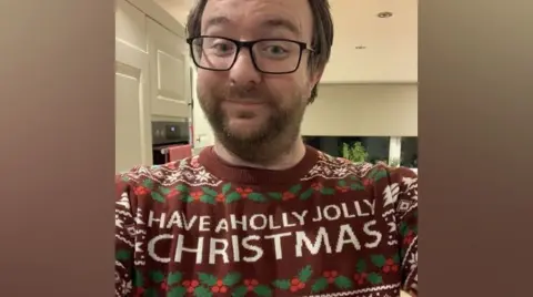 Dan Hiorns wearing a Christmas jumper, which has a pattern of holly leaves and berries, and shows the words "have a holly jolly Christmas". There are also white showflakes and Christmas trees on the jumper, which has a red background. Mr Hiorns is wearing glasses and smiling at the camera with his head slightly tilted.