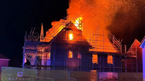  Peterborough Volunteer Fire Brigade (PVFB) A street view of a house with scaffolding outside with orange flames and plumes of orange smoke coming out of it.
