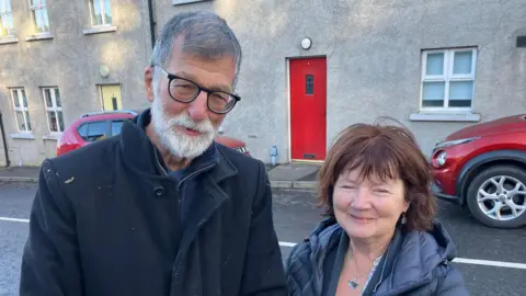 Vincent and Kate are standing side-by-side. Vincent has short grey hair and a white beard and moustache. He is wearing a black coat with a navy zip-up fleece and top. Kate has shoulder length brown hair and is wearing a navy coat and cardigan. Behind them is two red cars parked outside grey buildings, one with a red door and the other with a yellow.