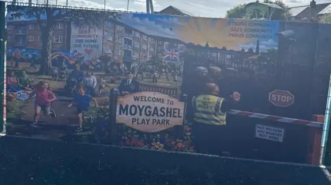 Banner on a play park fence, showing children playing sunshine and men being rejected at a barrier. 
