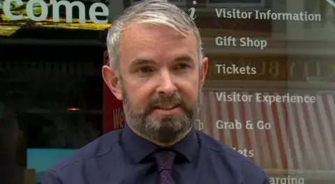 Visit Derry chief executive Odhran Dunne has short hair and a beard. He is standing in front of the Visit Derry office in the city centre and is wearing a navy shirt and purple tie