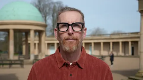 Shaun Whitmore/BBC Iwan Roberts standing in Eaton Park in Norwich. He is wearing a rust coloured shirt and is standing looking directly at the camera.