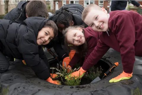 Julie Broadfoot A group of schoolchildren smiling while putting plants into soil while wearing orange gardening gloves. 