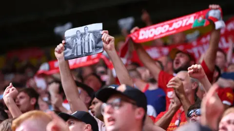 Reuters A Liverpool fan holds up a black and white image of footballers Diogo Jota and Andre Silva in a crowd of other supporters, many of whom are wearing red Liverpool football shirts. In the background, another fan holds up a red and white scarf bearing the name of Diogo Jota