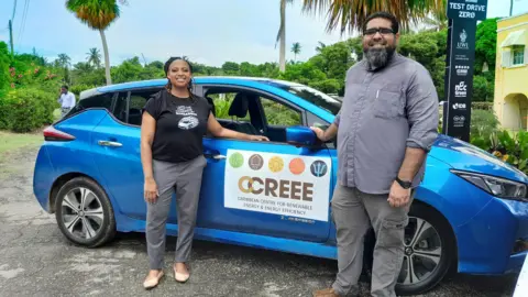 Dr Legena Henry berdiri di samping mobil biru dengan tanda bertuliskan "Pusat Energi Terbarukan dan Efisiensi Energi Karibia" di pintunya. 