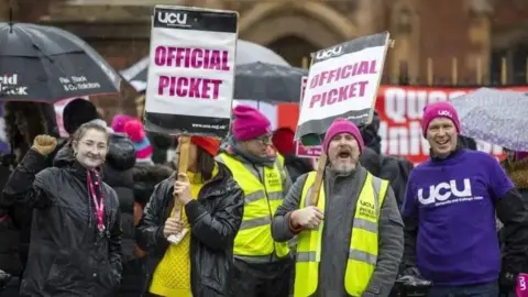 Union members, two of them in yellow high-viz vests on a picket line during previous strike action. Two of them are holding signs saying 'official picket' while a third is wearing a blue UCU top.