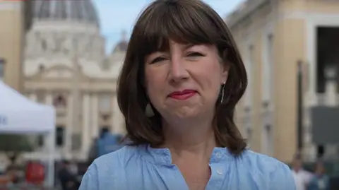 Professor Anna Rowlands, in a blue shirt, is standing in front of St Peter's square in Vatican City, with the huge white dome of St Peter's Basilica in the background.
