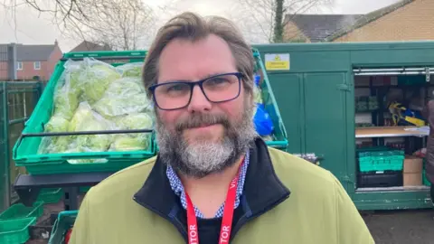 A man with short brown hair and a beard with grey patches, stands in front of food pantry stalls in a primary school playground. He wears glasses, a green jacket and a white and blue shirt, with a red lanyard.