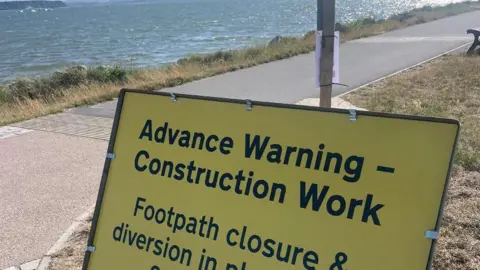 A yellow temporary roadworks sign which reads: 'Advance Warning - Construction Work. Footpath closure and diversion.' In the background is the riverside path and the water of Parkstone Bay in Poole Harbour.