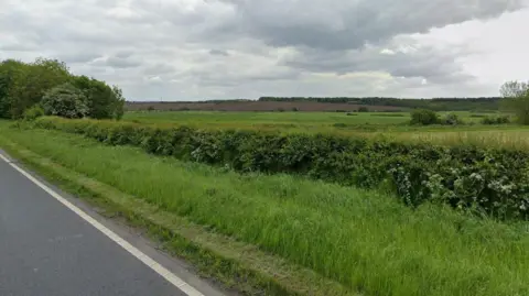 Land south of the A635 in South Yorkshire, near where four warehouses are planned. Beyond a hedge is green space and farm land, with a cloudy sky above. 