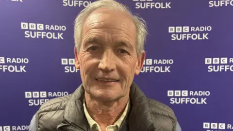 Harvey Bell/BBC George Burley wearing a black padded vest with a blue zip-up jumper and a checked shirt underneath, standing in front of a BBC Radio Suffolk backdrop.