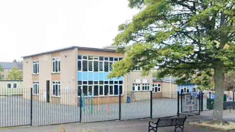 Google A brown square building with lots of rectangular windows behind a tall black gate. A street with a black metal bench and tall tree can also be seen
