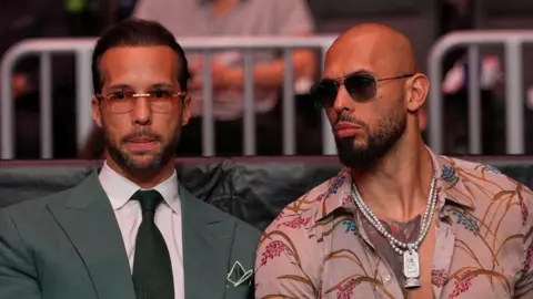 Julia Demaree Nikhinson/Pool via REUTERS The Tate brothers sit side by side facing forward. Both wear sunglasses. Tristan is wearing a suit, Andrew is wearing a floral shirt open at the collar with a large pendant necklace showing on his chest.
