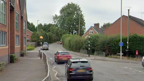 Google Streetview image of cars on the A529 in Market Drayton. Railings on the side of the path on the left lead to a zebra crossing, with hedges bordering the path on the right