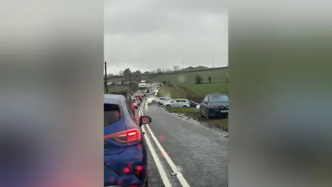 Ingrid Graham A long line of traffic along a rural road. More traffic is queuing from a junction. The sky is grey. It's been raining. 