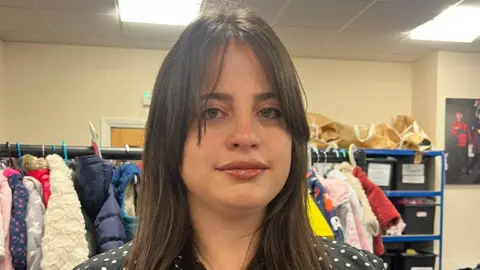 Volunteer Elsaida Golemi. She has long straight brown hair with a long overgrown fringe, parted in the middle. She is wearing pink lip gloss and a polka dot blouse. There is a rail of children's coats behind her. 