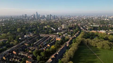 Getty Images An aerial, wide-angle view over leafy North London terrace housing, looking toward the distant City of London skyline featuring The Shard. Lush green parks and mature trees fill the foreground and middle ground under a clear, soft-lit sky.