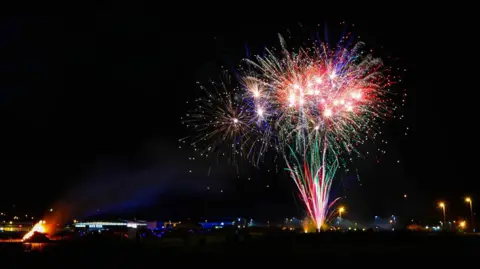 Colourful fireworks against a dark night sky. A bonfire burns in the bottom left corner.