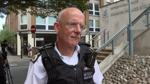 A Metropolitan Police officer speaking outside Holborn Police Station while wearing a police uniform and body armour.