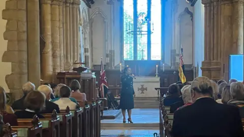 Hannah Gray/BBC The inside of a church with the pews full and people seen from the back. A stained glass window is seen at the back of the church and and a woman dressed in a dark blue suit is standing and singing in to a microphone.