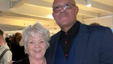 Ray Quinn A woman with short light blonde hair who wears a sparkly black dress, poses with Louis Emerick, who is taller, wears glasses and a blue suit. 