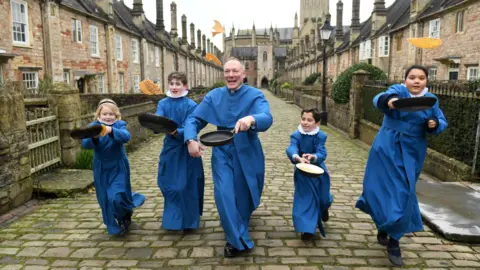 Four young choristers in blue robes attempt to flip pancakes as they run along a cobbled street. A member of the clergy, also dressed in blue, is in the middle of them. They are on Vicar's Close which is a traditional street lined with quaint cottages. The Cathedral is visible in the background.
