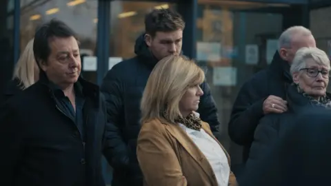 The Garden Productions A group of three men and two women outside a court, including on the far right Una Crown's niece Judy Payne. The men and Judy Payne are wearing dark clothes; a blonde woman in the front is wearing a tan jacket over a white shirt. 