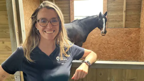 Mariachiara Apruzzese smiling wearing a navy blue Horses for Wellbeing polo shirt leaning on a stable door with her favourite horse, "Faith", in the background. The horse is dark brown with a white marking on its face.