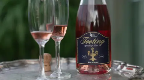 An open bottle of Tooting Sparkling Rosé on a silver tray. Next to it are two glasses with varying levels of wine in each glass, and a cork from the bottle.
