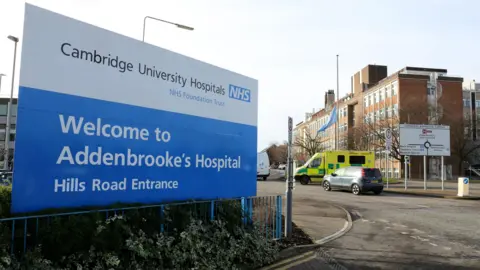 PA Media A large sign is on the left and is a welcome sign for Addenbrooke's Hospital. On the right is part of the hospital building and an ambulance and a car are on a road.