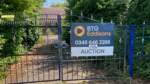 A set of blue metal gates stands at the end of a driveway. Behind them a single-storey building is visible. The gates are locked. On the right hand gate is a sign saying BTG Eddisons Auction.