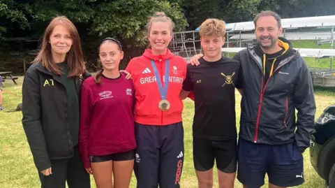 Group photo of five people next to each other. Middle person is wearing Team GB red hoodie and bronze medal around her neck.