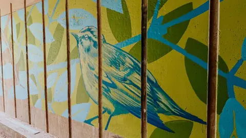 The mural has a large blue and yellow bird in its centre surrounded by blue tree branches and green leaves.
